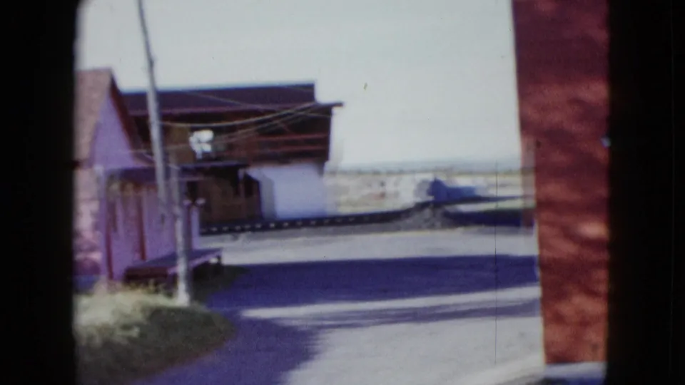 1958 Las Vegas home-movie frame with a paved road, utility pole, shaded building frontage, and a parked car across the street.