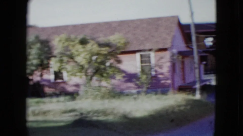 1958 Las Vegas frame showing a small house or outbuilding with grass, trees, utility pole, and part of a parked car at the edge.