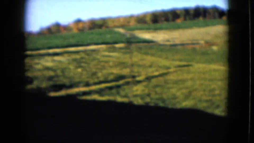 1958 home-movie frame looking across open grass toward a distant treeline, with heavy shadow from the viewing position.