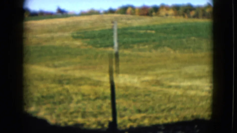 1958 8mm frame of a fence post in a green field, with rolling grassland and trees beyond in the reported Las Vegas area.
