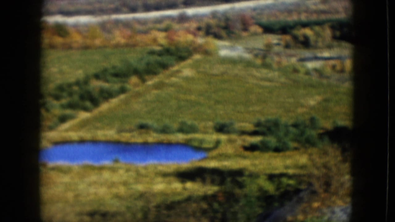 1958 8mm frame showing a small blue pond amid green rural fields and tree-lined terrain near the Las Vegas-labeled clip location.