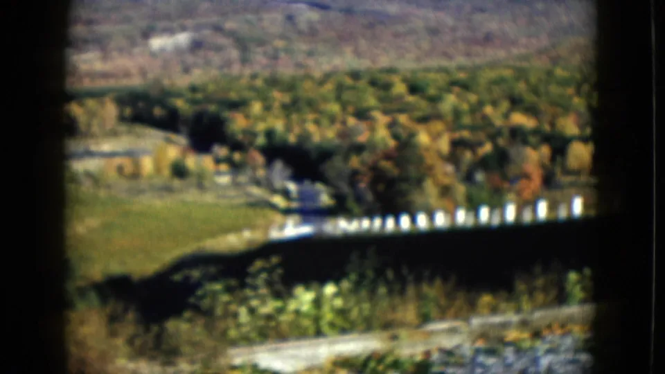 1958 8mm home-movie frame with white fence posts, green fields, trees, and distant hillside from a moving-vehicle viewpoint.
