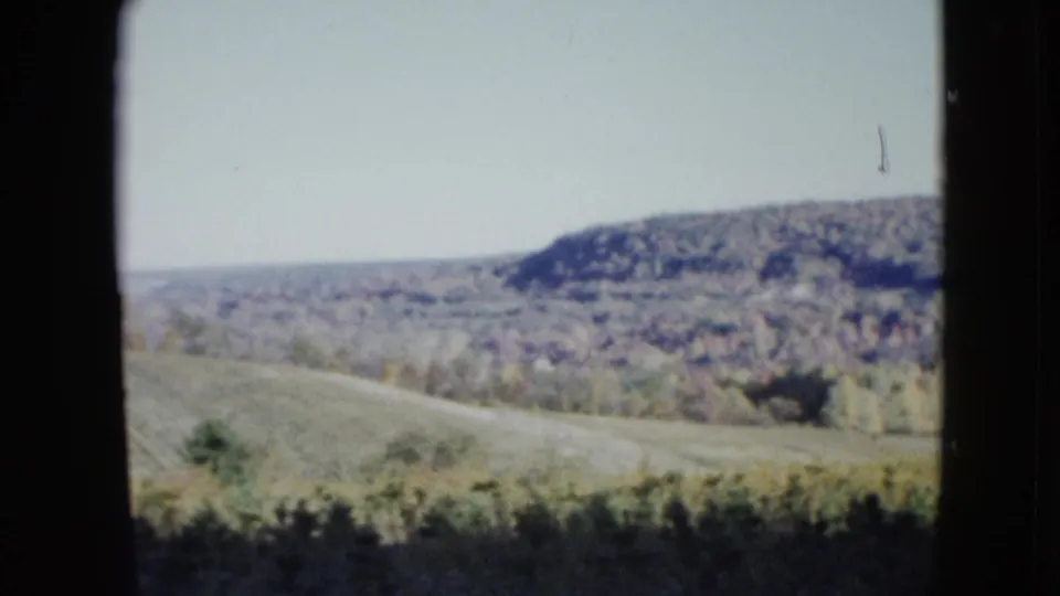 1958 8mm home-movie frame showing pale rolling countryside and open fields, viewed through a dark vertical window-like edge.