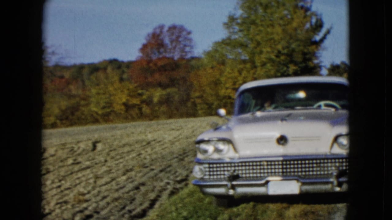 1958 8mm frame showing a chrome-front mid-century American car beside a cultivated field and autumn trees in rural landscape.