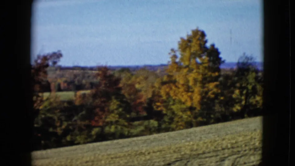 1958 8mm frame of a sloping rural field, autumn-colored trees, and distant hills, with no people visible.