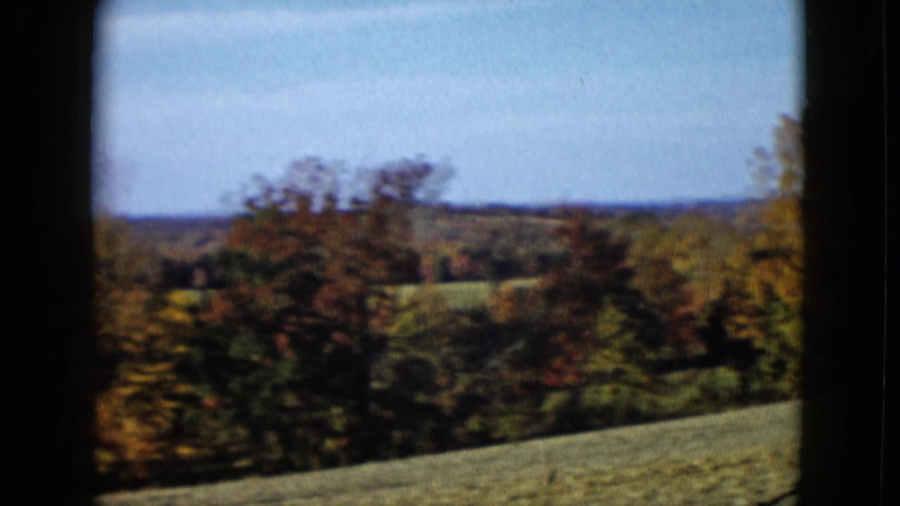 1958 8mm home-movie view of autumn trees, green fields, and distant hills framed by a dark window edge.