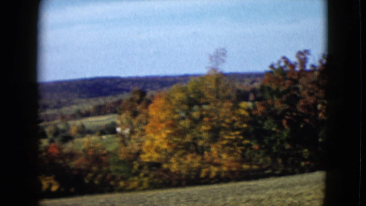 1958 8mm frame of colorful fall trees and rolling fields, with a tiny light structure in the distance.