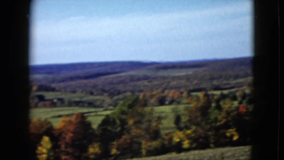 Soft 8mm view across a broad rural valley, with fall trees in front and layered hills beyond.