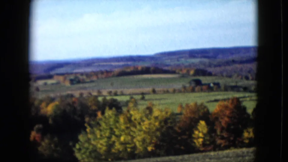1958 home-movie frame showing patchwork fields, wooded ridges, and autumn foliage from a moving vehicle.