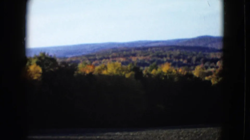 Soft 1958 travel footage frame showing fall-colored trees and distant ridges across a dark foreground band.