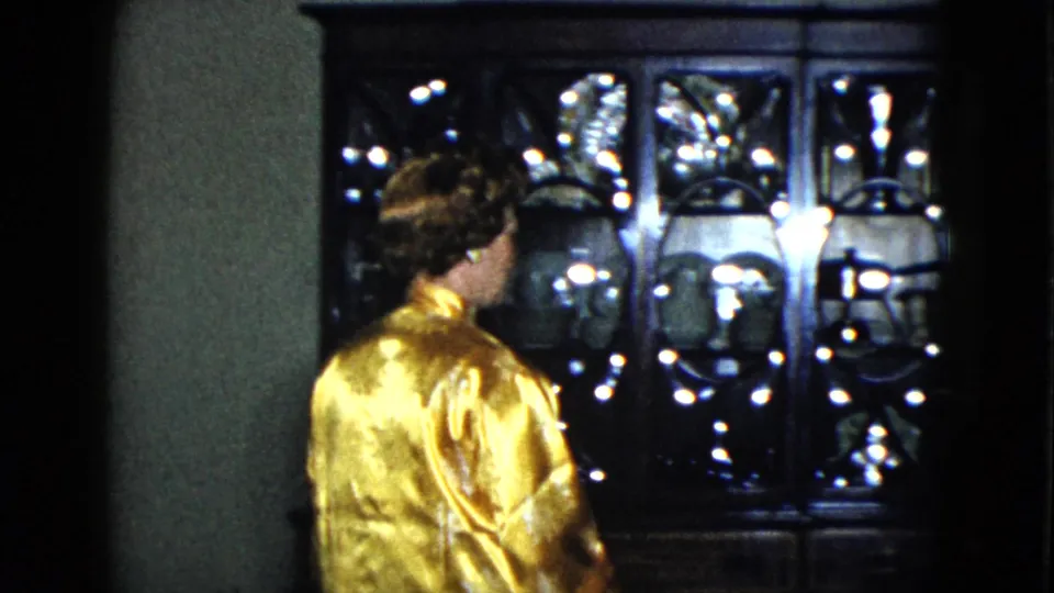 In a 1958 Las Vegas interior, a woman in a shiny gold robe faces a dark decorative glass cabinet.