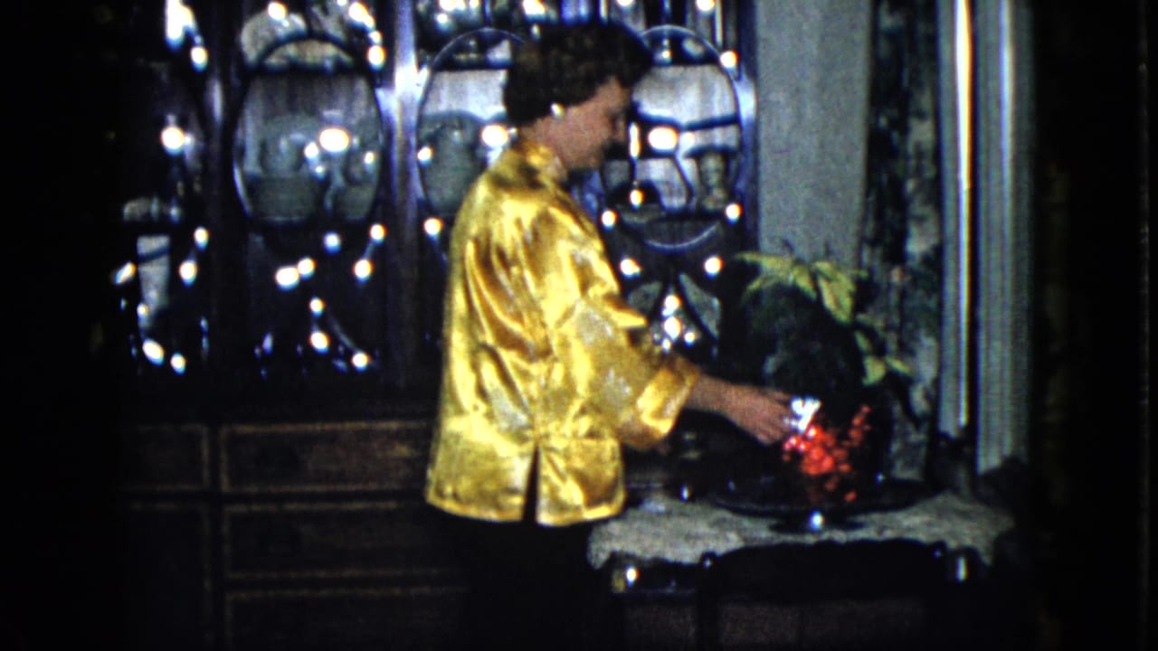 Woman in a gold satin jacket arranges red flowers beside a dark glass cabinet.