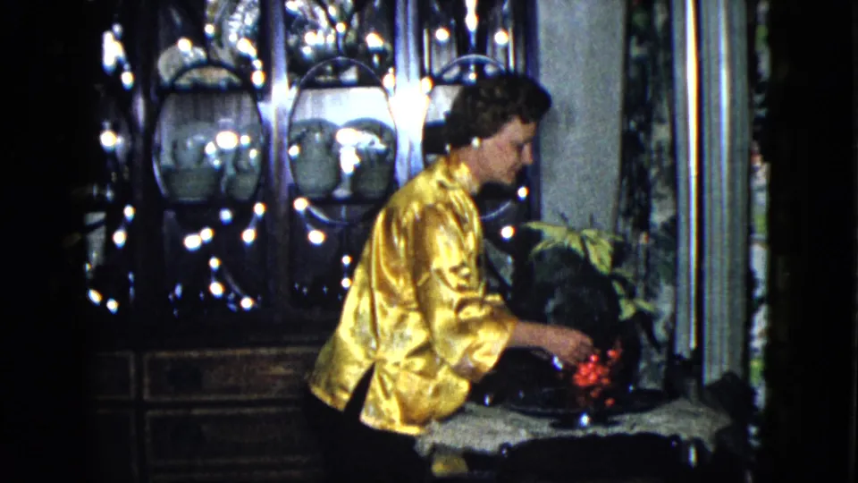 The woman leans in to arrange red flowers, with glassware and dishes visible in a dark cabinet behind her.