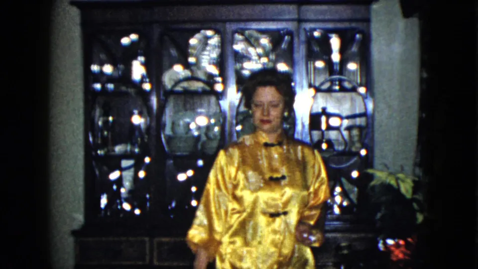 In the Las Vegas residence, the woman faces the camera in her gold jacket with the flower arrangement partly visible beside her.