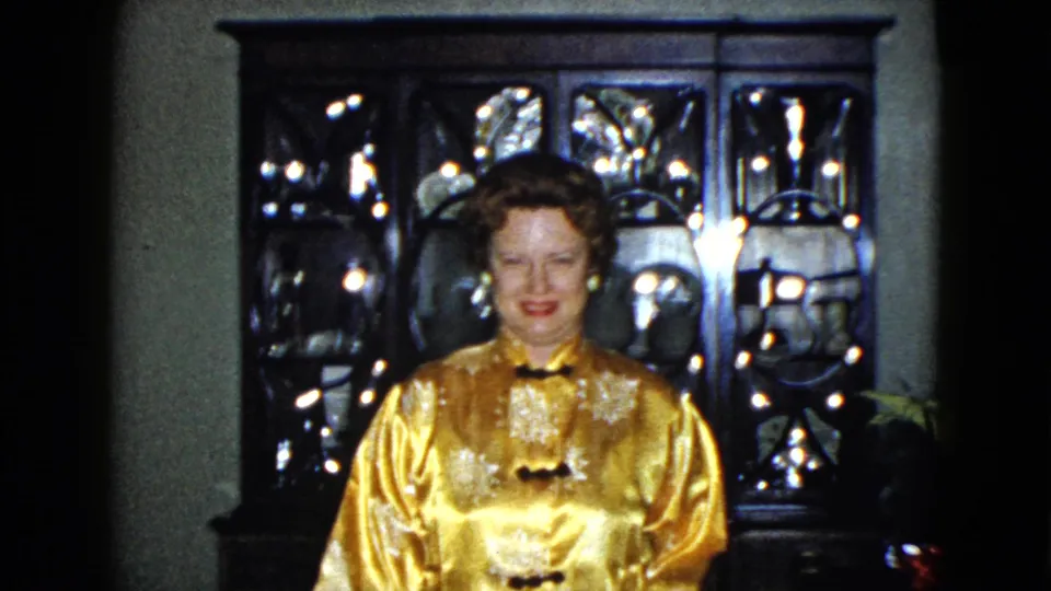 The woman smiles toward the camera in a 1958 Las Vegas interior, standing before a lit glass cabinet and nearby flowers.