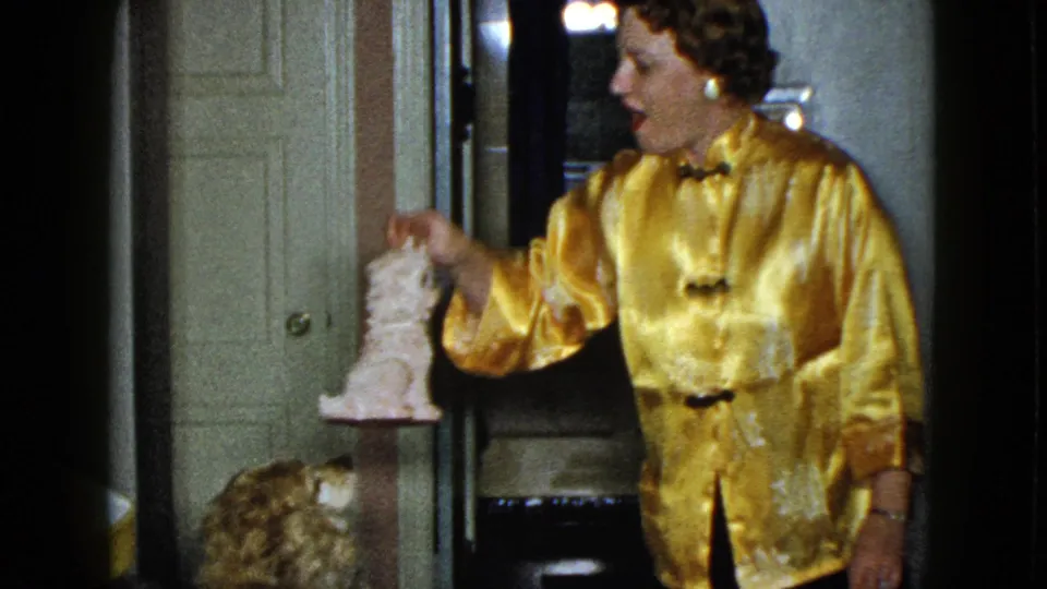 A woman in a shiny gold jacket smiles down at a fluffy dog while holding a white stuffed toy in this 1958 home-movie frame.