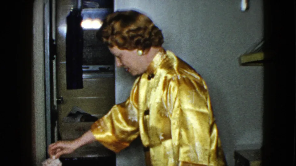 A woman in a gold satin jacket leans across the room near a plaid sofa, with a door and dark interior furnishings behind her.