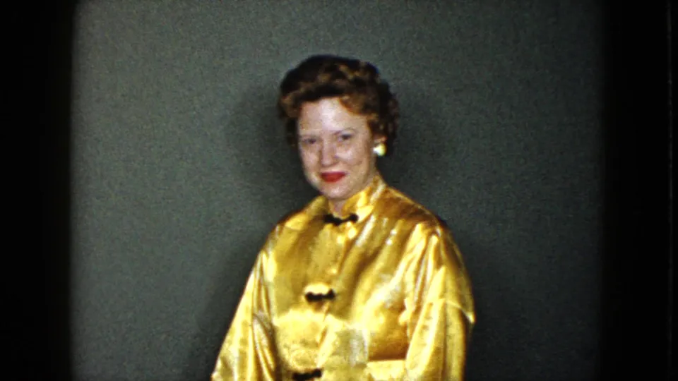 A woman in a shiny gold dress sits in the Las Vegas home interior and faces the home-movie camera.