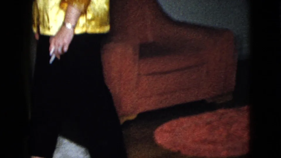 The camera frames the woman's gold blouse, dark pants, cigarette, and a red upholstered chair in the private interior.