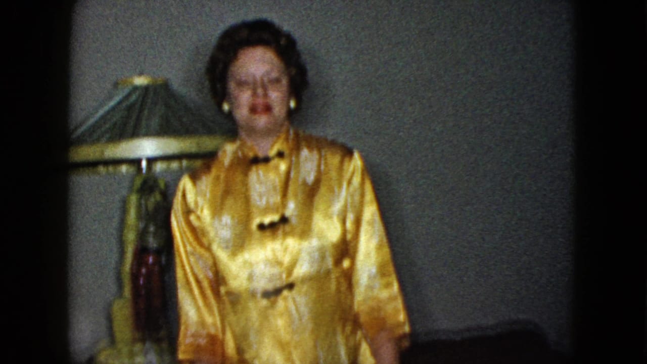 Woman in a shiny gold dress standing indoors beside a shaded lamp.