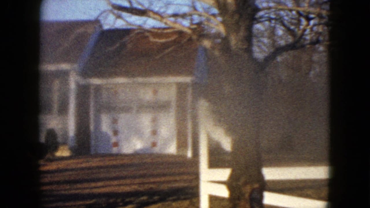 1958 Las Vegas home-movie frame showing a white residential house with a brown roof, bare tree branches, deep yard shadows, and a pale fence in the foreground.