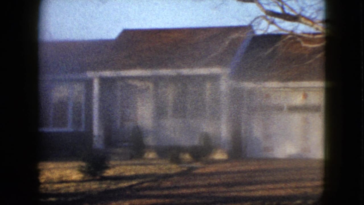 1958 Las Vegas exterior frame showing a white-sided suburban house with a brown roof, bare tree limbs overhead, and low bushes along the front yard.