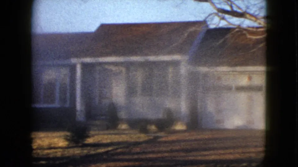 1958 Las Vegas exterior frame showing a white-sided suburban house with a brown roof, bare tree limbs overhead, and low bushes along the front yard.