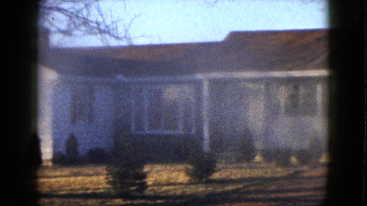 Soft 8mm frame of a white Las Vegas house exterior in 1958, with front windows, low shrubs, bare branches, and strong shadows crossing the yard.