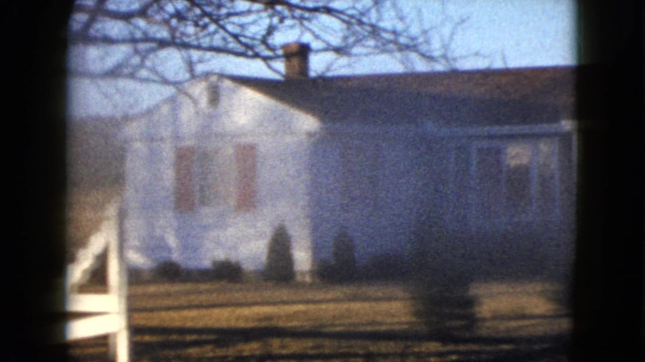 1958 Las Vegas home-movie frame showing the side or front of a white house with chimney, red shutters, bare branches, lawn, shrubs, and a pale fence edge.