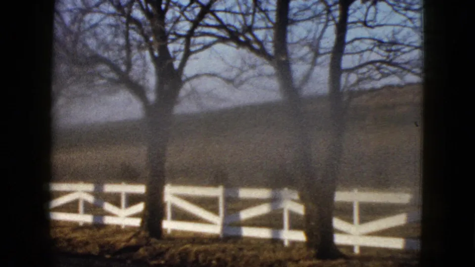 The final frame shows two leafless trees and a white rail fence across a quiet rural property in grainy 8mm color.