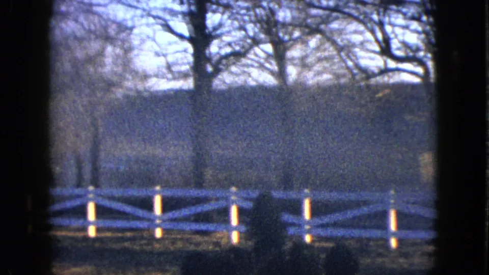 Soft 1958 8mm frame from the Las Vegas metadata sequence with white fencing, bare trees, and a dim wooded backdrop.
