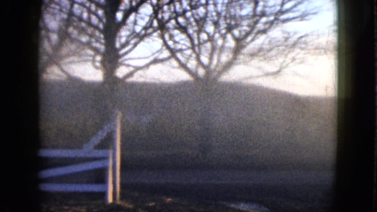 1958 8mm frame showing bare trees, a white rail fence, muted grass, and hazy low hills through a dark foreground frame.