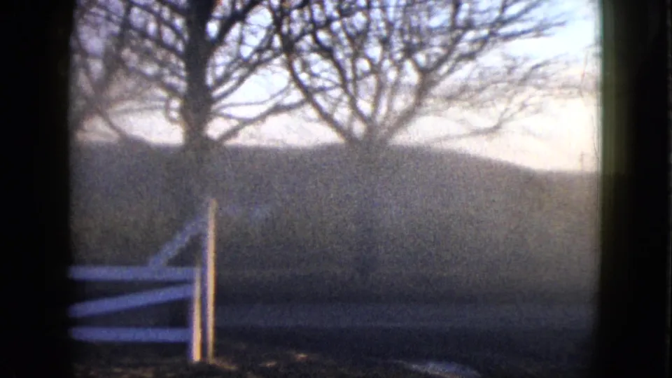 1958 8mm frame showing bare trees, a white rail fence, muted grass, and hazy low hills through a dark foreground frame.
