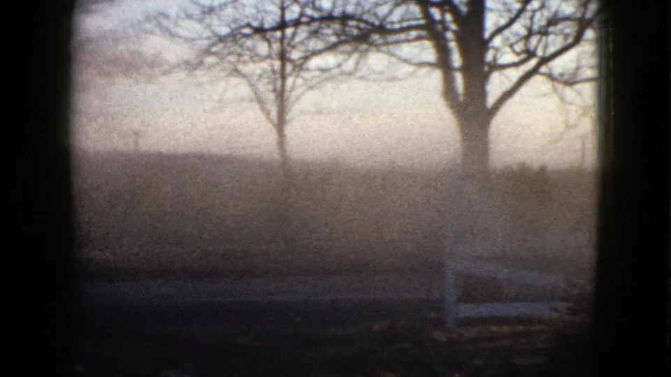 1958 8mm frame showing leafless trees, a white wooden fence, dark foreground grass, and a washed, hazy sky.