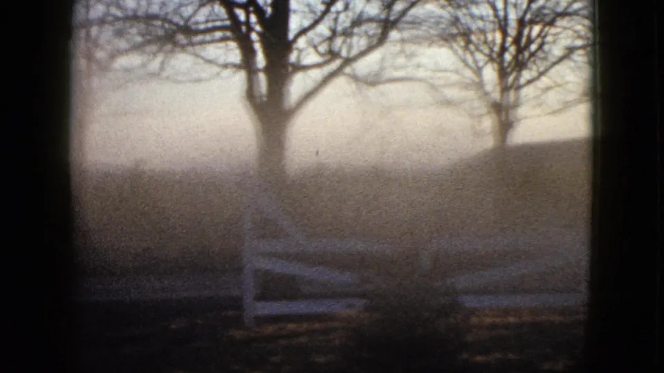 1958 8mm frame showing a white fence crossing a dim rural foreground with bare trees and a softly lit hillside behind it.