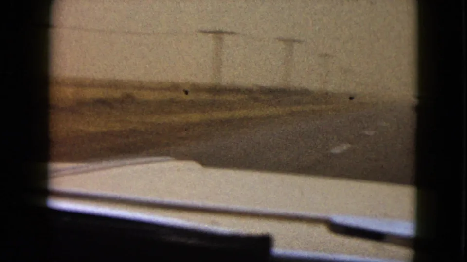 Soft, grainy 8mm frame from inside a moving car, showing the road edge, hood, and telephone poles fading into fog.