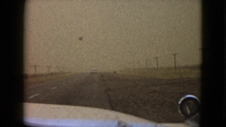 1958 home-movie road view near Las Vegas, looking over the car hood toward a foggy two-lane road lined with telephone poles.