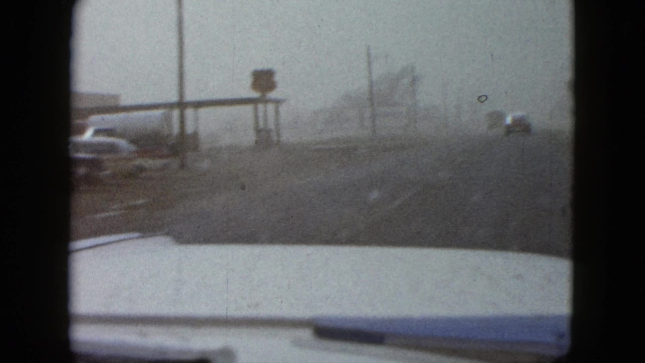 Car hood foreground facing a dusty road with roadside posts, signs, and distant vehicles.
