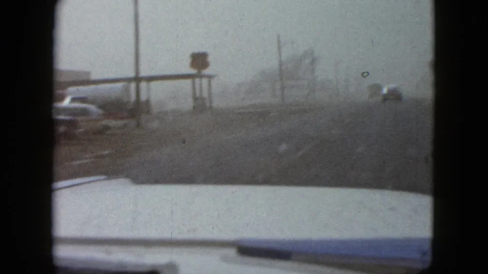 1958 Las Vegas street view from a moving car, with poor visibility, roadside signs, utility poles, and vintage vehicles ahead.