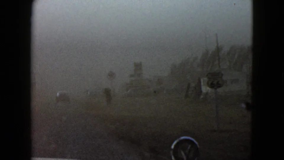 1958 Las Vegas 8mm frame showing a windshield view toward a dusty roadside with signposts, bare trees, and low structures.