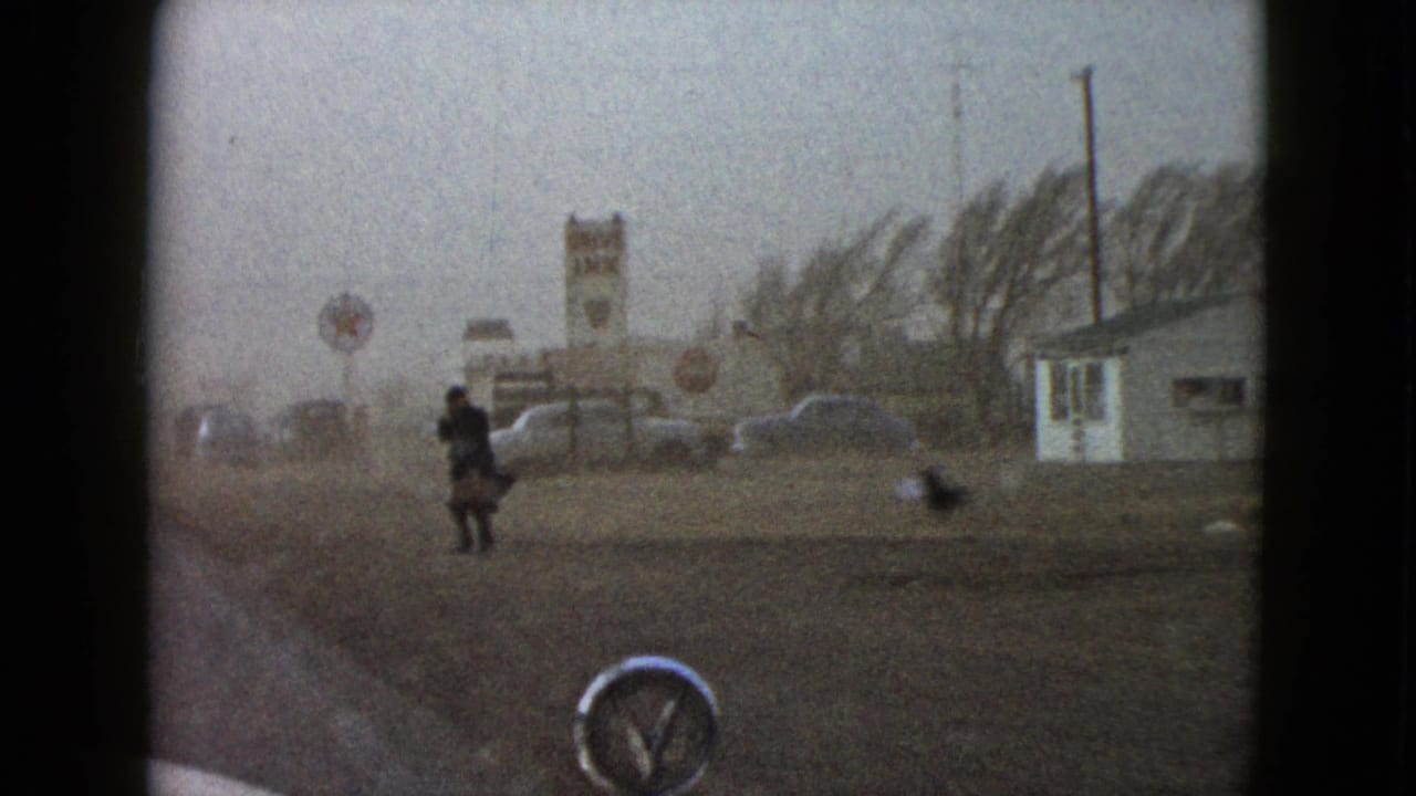 1958 Las Vegas-area 8mm frame showing a lone roadside man, small structures, a car hood, and a gray windy sky.