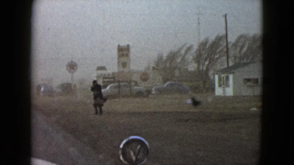 1958 Las Vegas-area 8mm frame showing a lone roadside man, small structures, a car hood, and a gray windy sky.