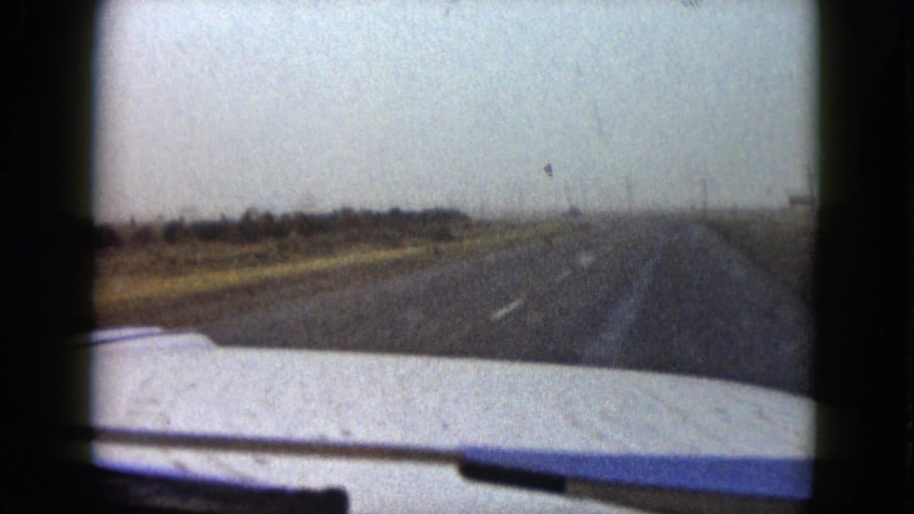 Passenger-view 8mm frame of a long rural road near Las Vegas, with car hood, open fields, and low horizon under gray sky.