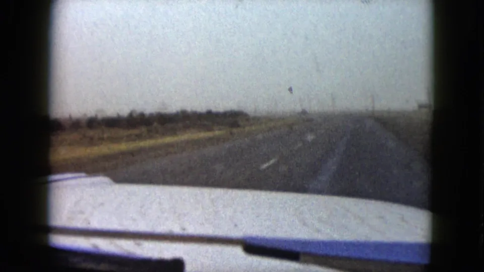 Passenger-view 8mm frame of a long rural road near Las Vegas, with car hood, open fields, and low horizon under gray sky.
