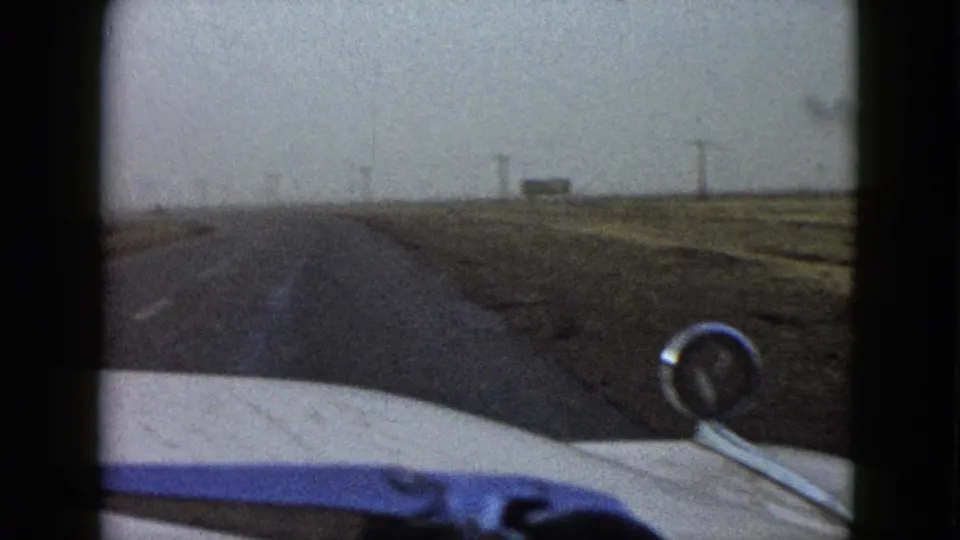 1958 8mm driving view with a white car hood, round mirror, straight country road, fields, and distant roadside signs.