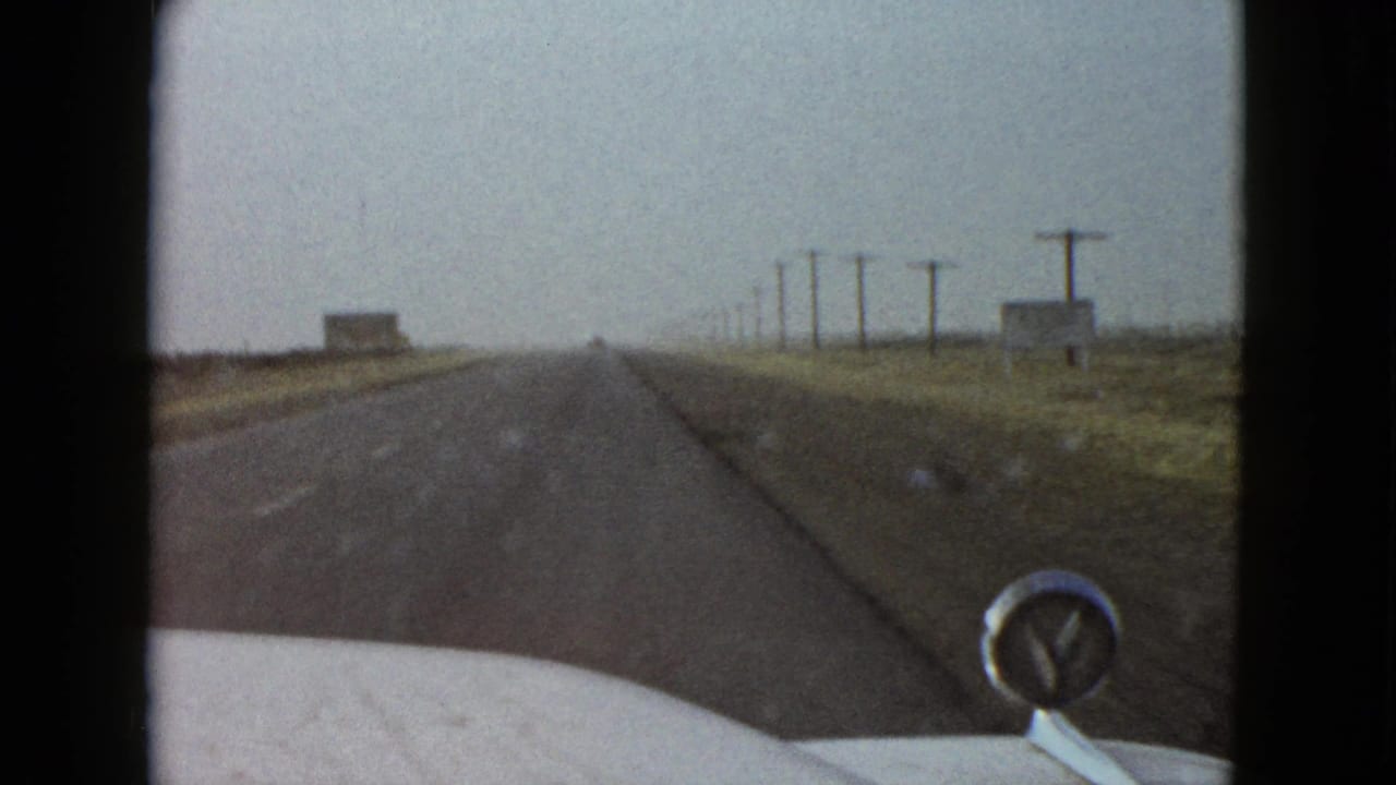 1958 Las Vegas-area home-movie frame from inside a car, showing an empty asphalt road, hood ornament, telephone poles, and sparse desert terrain.