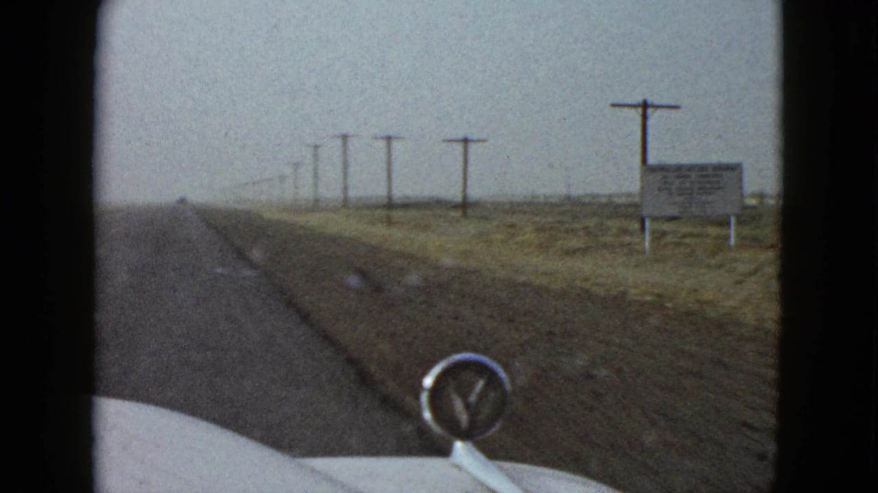 1958 Las Vegas-area windshield view with a roadside sign, wooden telephone poles, arid brushland, and the car hood ornament in the foreground.