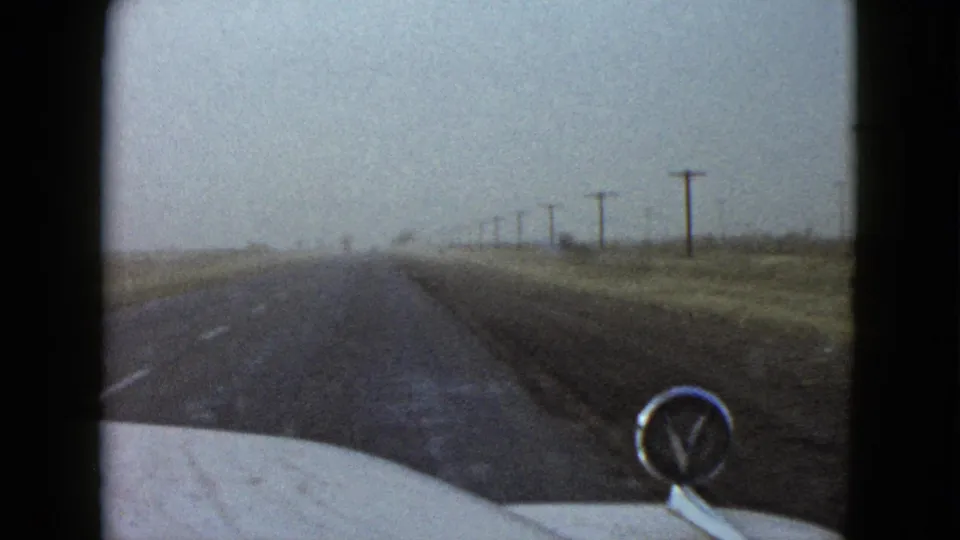 Las Vegas-area 1958 road footage showing a forward windshield view, soft haze, telephone poles, dry roadside brush, and a visible car hood ornament.
