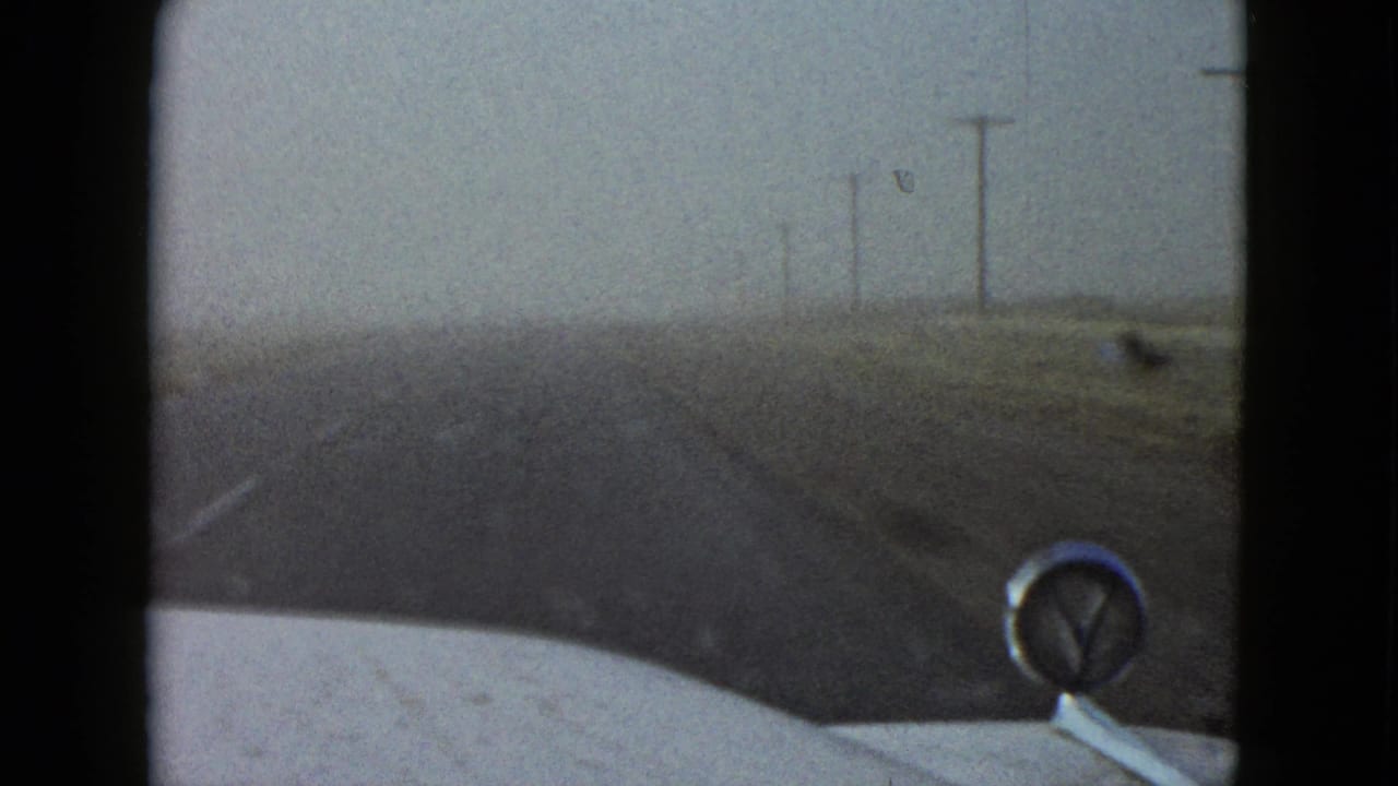 1958 Las Vegas-area home-movie frame from inside a moving car, showing a straight road, dry roadside, and chrome hood ornament.
