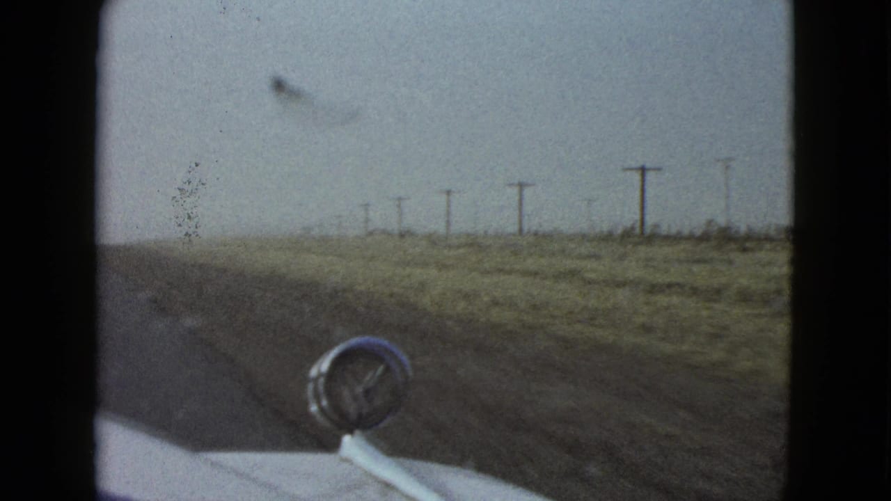 1958 Las Vegas-area home-movie frame looking from a moving car past a chrome hood ornament toward a flat roadside with utility poles.